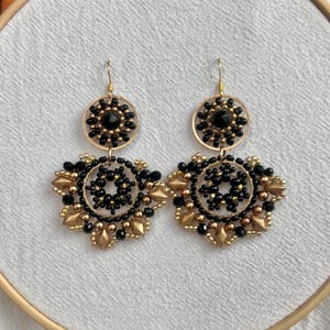 May include: A pair of gold and black beaded earrings with a circular design. The earrings feature multiple layers of beads, including small black beads and larger gold beads.
