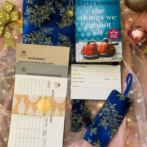 Puede incluir: Una colección de accesorios de lectura, que incluye un libro, una funda de libro floral azul, un marcador y un juego de desafío de lectura. La portada del libro presenta un par de zapatos rojos. Los artículos están dispuestos sobre una superficie rosa.
