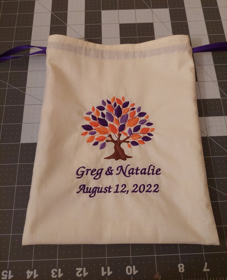 May include: A white drawstring bag with an embroidered tree design in shades of orange, purple, and brown. The bag has a purple drawstring and the text "Greg & Natalie August 12, 2022" is embroidered below the tree.