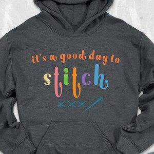 May include: A dark gray hooded sweatshirt with the text "It's a good day to stitch" in rainbow colors. The text is in a playful, handwritten font.