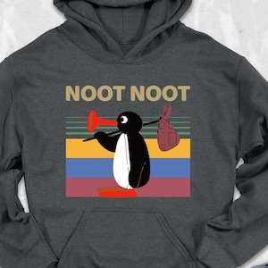 May include: Dark gray hoodie with a penguin graphic holding a red megaphone and a brown glove, with the text "NOOT NOOT". The penguin is set against a colorful striped background. The hoodie has a hood and a kangaroo pocket.