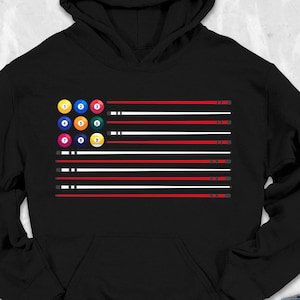 May include: Black hoodie with a graphic design of a billiards-themed flag. The flag design features a cluster of billiard balls in the upper left corner, with red, white, and black stripes. The hoodie is surrounded by accessories like sunglasses, shoes, and a backpack.
