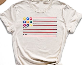 May include: A beige t-shirt with a graphic of a pool table flag. The flag has red and white stripes with a circle of billiard balls in the top left corner.