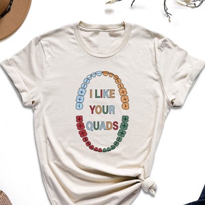 May include: A beige t-shirt with a graphic of a set of teeth with the text "I like your quads" printed on it.