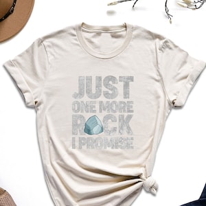 May include: A beige t-shirt with the text "JUST ONE MORE ROCK I PROMISE" in a glittery font. A small blue rock graphic is included in the text.