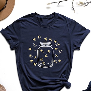 May include: A navy blue t-shirt with a white outline of a mason jar filled with fireflies. The fireflies are yellow and there are small white stars and diamonds around the jar.