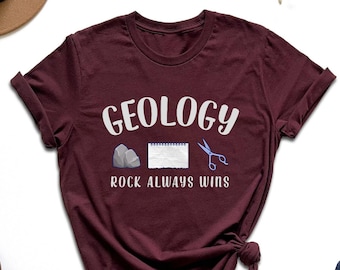 Könnte beinhalten: Burgunderfarbenes T-Shirt mit weißem Text, der "Geology Rock Always Wins" lautet. Das Shirt zeigt eine kleine Grafik von Stein, Papier und Schere.