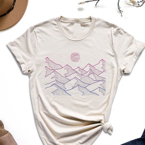 May include: A beige t-shirt with a graphic of a mountain range with a sun in the background. The mountains are outlined in pink and blue.