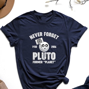 May include: A navy blue t-shirt with a white graphic that says "Never Forget Pluto, Former "Planet"" with the years 1930 and 2006 on either side of the graphic. The graphic also includes a cartoon of Pluto with a sign that says "Dwarf Planets Matter".