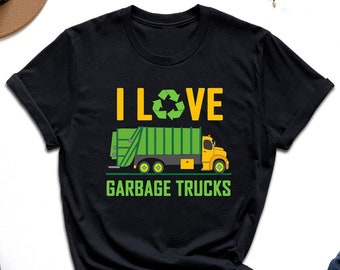 May include: Black t-shirt with the text "I LOVE GARBAGE TRUCKS" in yellow and green. The design includes a green recycling symbol and a yellow and green garbage truck illustration. The shirt is displayed with jeans and accessories.