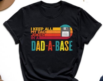 May include: Black t-shirt with the text "I KEEP ALL MY DAD JOKES IN A DAD-A-BASE" in a retro design. The design includes a floppy disk graphic and a sunset-style color scheme. The shirt is paired with blue jeans and other accessories.