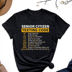May include: Black t-shirt with a white graphic that says "Senior Citizen Texting Code" and lists common texting abbreviations and their meanings for older adults.