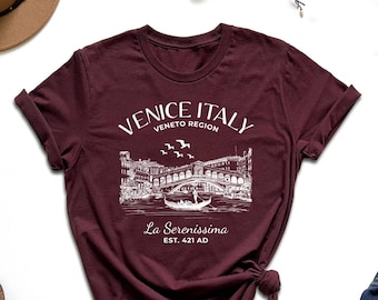May include: A maroon t-shirt with a white graphic design of a bridge in Venice, Italy. The text on the shirt reads "Venice Italy Veneto Region La Serenissima Est. 421 AD".