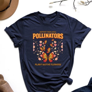 May include: A navy blue t-shirt with a graphic of butterflies and flowers. The text on the shirt reads "Support Your Local Pollinators Plant Native Flowers".