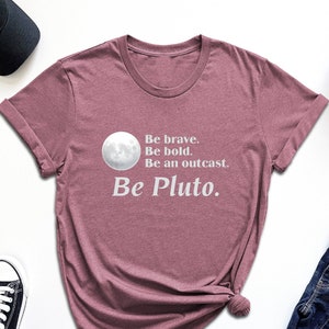 May include: Mauve t-shirt with the text "Be brave. Be bold. Be an outcast. Be Pluto." A graphic of a full moon is above the text. The shirt is styled with black sunglasses, sneakers, a black cap, and jeans.