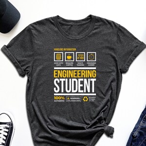 May include: A dark grey t-shirt with a white graphic that says "Engineering Student" and includes a list of requirements for the job, such as "Good with Math", "Requires Coffee", "Multi-tasking", and "Cramming Expert".