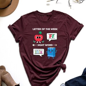 May include: A maroon t-shirt with the text "Letter of the Week" and "Sight Word" printed on it. The shirt features a cartoon apple with the letter "F" and a cartoon book with the word "this" printed on it.