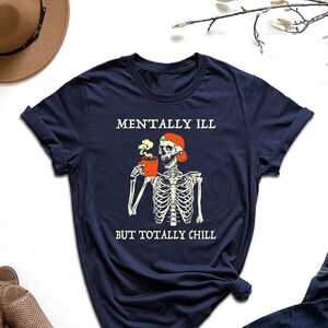 May include: Navy blue t-shirt with a skeleton wearing a Santa hat and holding a cup of coffee. The text "Mentally Ill But Totally Chill" is printed on the shirt.