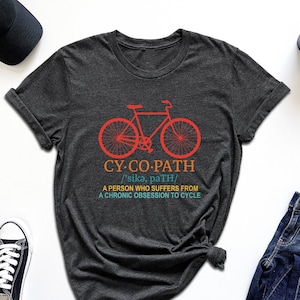 May include: A dark gray t-shirt with a red bicycle graphic and the text "CY-CO-PATH /'sika, paTH/ A PERSON WHO SUFFERS FROM A CHRONIC OBSESSION TO CYCLE"