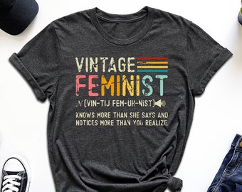 May include: A dark grey t-shirt with the text "VINTAGE FEMINIST (VIN-TIJ FEM-UH-NIST)" in colorful letters. The text is followed by "KNOWS MORE THAN SHE SAYS AND NOTICES MORE THAN YOU REALIZE".