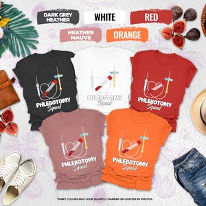 May include: A collection of crew neck t-shirts in various colors, including dark grey, white, red, orange, and heather mauve. Each shirt features a medical-themed graphic with the text "PHLEBOTOMY Squad". The shirts are displayed with accessories like shoes, a hat, and figs.