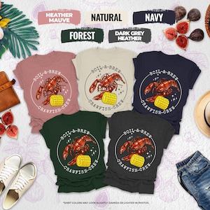 May include: A collection of t-shirts in various colors, including heather mauve, natural, navy, forest, and dark grey heather. Each shirt features a circular graphic with a crawfish and corn, with the text "BOIL-&-BREW CRAWFISH-CREW".