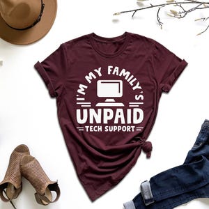 May include: A maroon short-sleeved t-shirt featuring a white graphic of a computer monitor and the text I'M MY FAMILY'S UNPAID TECH SUPPORT. The shirt has a crew neckline and is displayed with a brown hat, blue jeans, and tan sandals.
