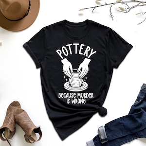 May include: Black t-shirt with the word "POTTERY" above a graphic of hands shaping a pot. Below the graphic, the text reads "BECAUSE MURDER IS WRONG". The shirt is displayed with a brown hat, shoes, and jeans.