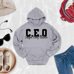 May include: A light gray hooded sweatshirt featuring the black text C.E.O. and the words Conquer Every Obstacle printed on the front. The garment includes a hood, long sleeves, and a front kangaroo pocket, presented as a central clothing item.