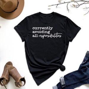 May include: Black t-shirt with the text "currently avoiding all responsibilities" in white script. The shirt is styled with blue jeans, a brown hat, and open-toe shoes. A starfish and greenery are also in the image.