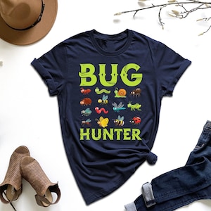 May include: Navy blue t-shirt with the words "BUG HUNTER" in green, featuring colorful illustrations of various insects. The shirt is styled with blue jeans, a brown hat, and sandals.