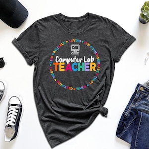 May include: Dark gray t-shirt with the words "Computer Lab TEACHER" in a colorful circular design. The shirt is surrounded by accessories, including sunglasses, sneakers, and a smartwatch. The shirt is made of a soft material.