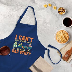 May include: A blue apron with a white pocket and the text "I can't talk right now I'm doing hot dad stuff" in orange. The apron has a graphic of a beer, tools, and a grill.