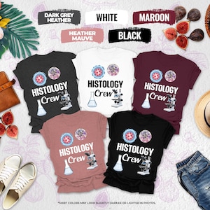 May include: A collection of t-shirts in various colors, including black, white, maroon, and heather mauve. Each shirt features the text "HISTOLOGY Crew" along with a graphic of cells, a beaker, and a microscope. The shirts are displayed with accessories like shoes, a hat, and figs.
