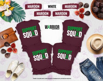 May include: A collection of maroon and white t-shirts with the words "SUPPORT SQUAD" and "WARRIOR" printed on them. The shirts have a green ribbon design, and the text "Awareness Palsy Awareness" is also printed on the shirts. The shirts are displayed with accessories.