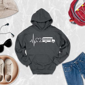 May include: A dark gray hooded sweatshirt featuring a white graphic print on the chest. The design displays a heartbeat line that transitions into a heart shape and then into a school bus silhouette. The hoodie has long sleeves and a front pocket.