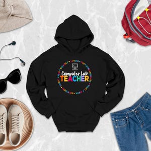 May include: A black hooded sweatshirt featuring a graphic design with the text Computer Lab Teacher in colorful letters, surrounded by a circular border of descriptive words like Loving, Fun, and Kind, and a small computer icon at the top.