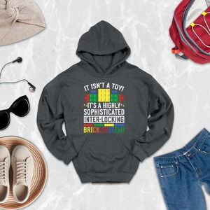 May include: A dark gray hooded sweatshirt featuring the white text IT ISN'T A TOY! IT'S A HIGHLY SOPHISTICATED INTER-LOCKING BRICK SYSTEM! with colorful yellow, green, red, and blue brick graphics printed on the center of the chest.