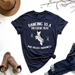 May include: Navy blue t-shirt with the text "Dancing to a Different Beat" and "Rare Disease Awareness". The shirt features a zebra graphic. The shirt is paired with blue jeans, a brown hat, and sandals.