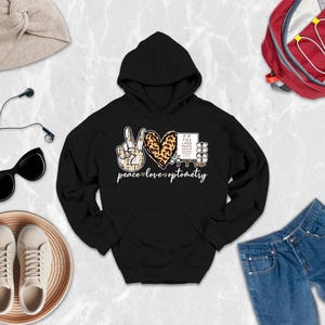 May include: A black hooded sweatshirt featuring a graphic design with a peace sign, a leopard print heart, and an eye chart. The text below the graphic reads peace love optometry. The design incorporates small heart icons between the words.