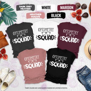 May include: A collection of Optometry Squad t-shirts in various colors, including dark grey heather, white, maroon, heather mauve, and black. Each shirt features the text "OPTOMETRY SQUAD" with eyeglasses graphic. The shirts are displayed with accessories.