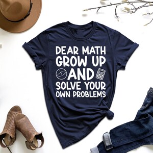 May include: Navy blue t-shirt with the text "DEAR MATH GROW UP AND SOLVE YOUR OWN PROBLEMS" in white. The shirt features a graphic of a sphere and a calculator. The shirt is paired with blue jeans, a brown hat, and sandals.