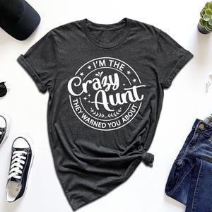 May include: A dark charcoal gray short-sleeve t-shirt featuring a white circular graphic with the text I'M THE Crazy Aunt THEY WARNED YOU ABOUT, surrounded by stars and leaf accents, displayed with a black baseball cap, sunglasses, sneakers, and jeans.