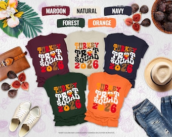May include: Five t-shirts in maroon, natural, navy, forest green, and orange, each featuring a graphic with the text Turkey Trot Squad 2026 and a cartoon turkey illustration. The shirts are displayed with a brown bag, figs, a hat, and sneakers.