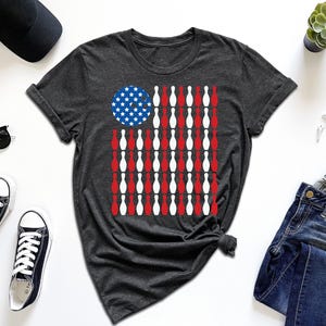 May include: Dark gray t-shirt featuring a graphic design of the American flag made of bowling pins. The flag design uses red and white pins for the stripes and a blue circle with white stars for the stars. The shirt is surrounded by accessories.