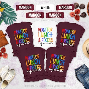 May include: A collection of t-shirts in maroon and white, each featuring the text "MONITOR LUNCH & RECESS squad" in colorful, rainbow-style lettering. The shirts are arranged with various accessories, including a brown leather bag, figs, and a straw hat.