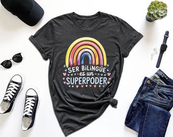 May include: Dark gray t-shirt with a watercolor rainbow design and the text "SER BILINGÜE es un SUPERPODER". The shirt is styled with black sunglasses, sneakers, a watch, and jeans.