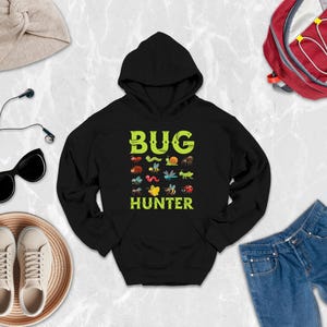May include: A black hooded sweatshirt featuring the text BUG HUNTER in lime green letters, accompanied by a grid of colorful insect and arachnid illustrations including a ladybug, caterpillar, snail, spider, dragonfly, bee, and butterfly.