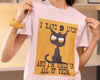 Op de afbeelding: Lichtroze T-shirt met een cartoon kat ontwerp. De zwarte kat met gele ogen heeft een humeurige uitdrukking. Het ontwerp staat op een gele achtergrond, met de tekst: "I HAVE 9 LIVES AND I'M TIRED IN ALL OF THEM."