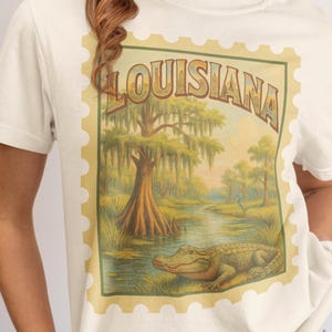 May include: Cream-colored t-shirt featuring a vintage-style Louisiana design. The design includes the word "LOUISIANA" in a retro font, a swamp scene with an alligator, trees, and a waterway, framed by a postage stamp border.